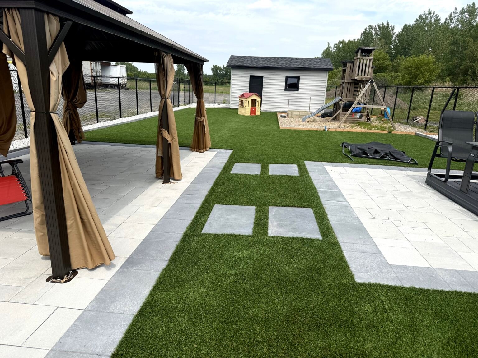 Cour familiale en gazon synthétique entre un patio de pavé, un gazebo et l'aire de jeux